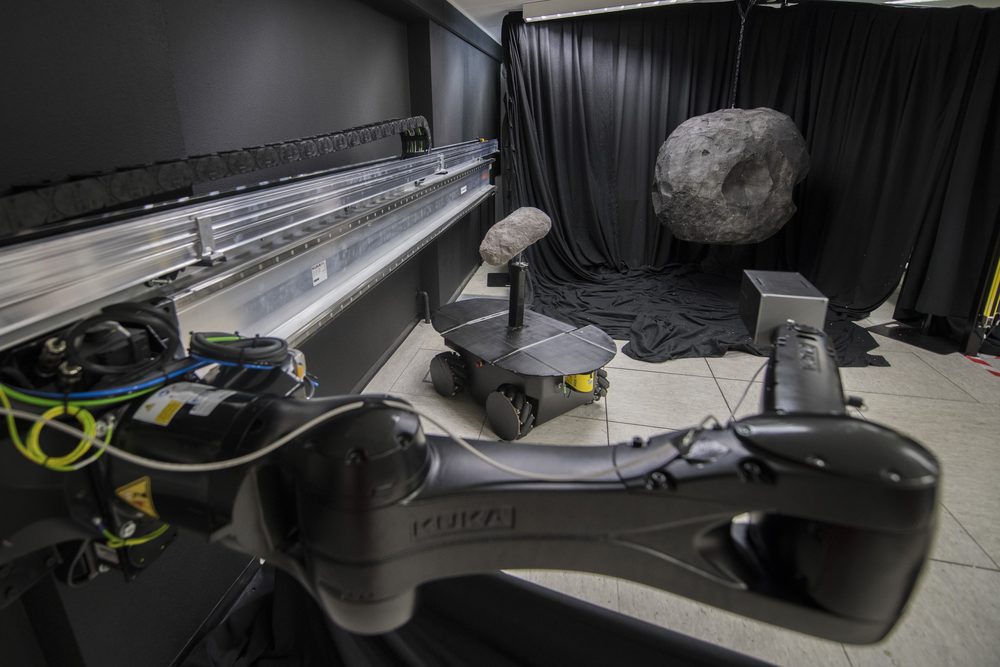 A black KUKA arm in the foreground, grey rock models against black drapes
