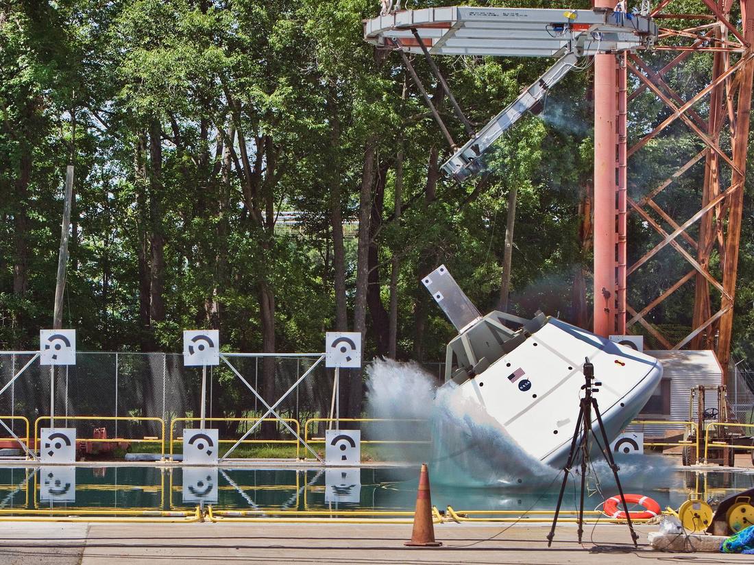 White capsule splashing into a basin beneath orange gantry legs, spray rising past photo reference targets