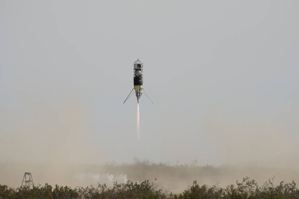 Slender rocket with splayed legs in flight over desert scrub, a thin plume beneath
