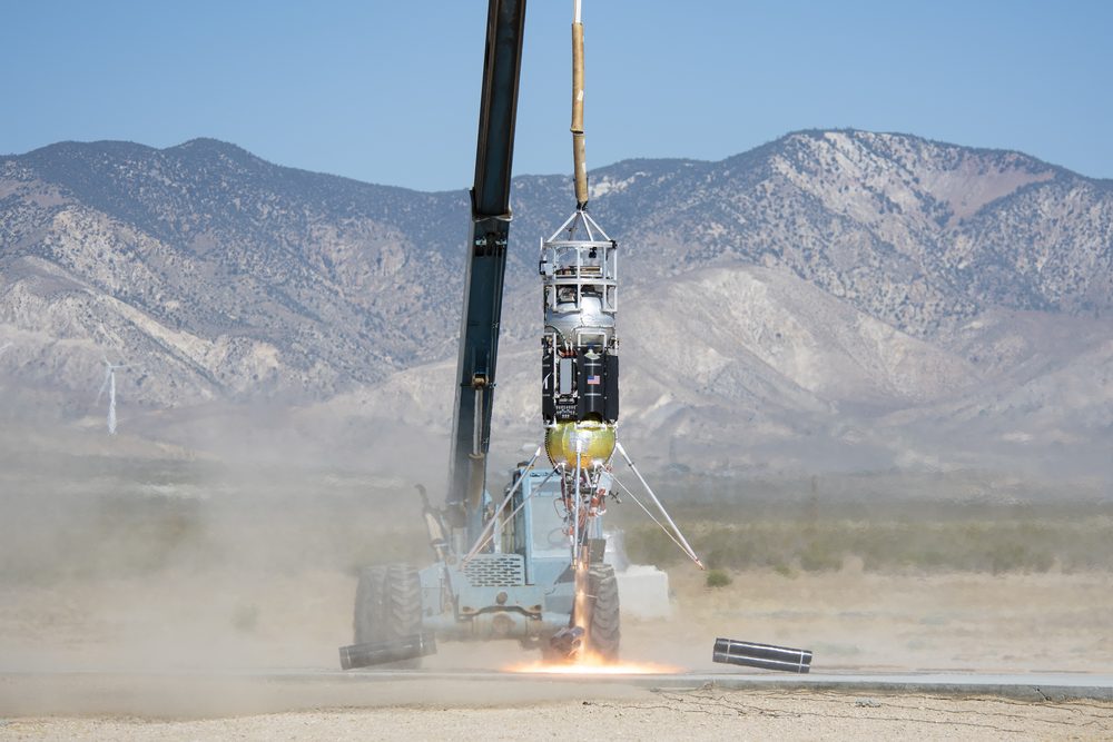 Slim rocket lifting on an orange flame amid dust, a crane cable overhead, mountains behind