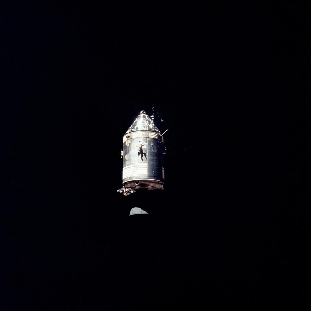 Sunlit cylindrical spacecraft with a conical nose, alone against black, grey surface below