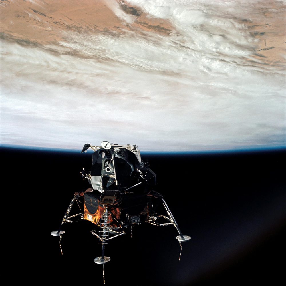 Four-legged lander in dark space, its pads extended, cloud-covered Earth filling the upper frame
