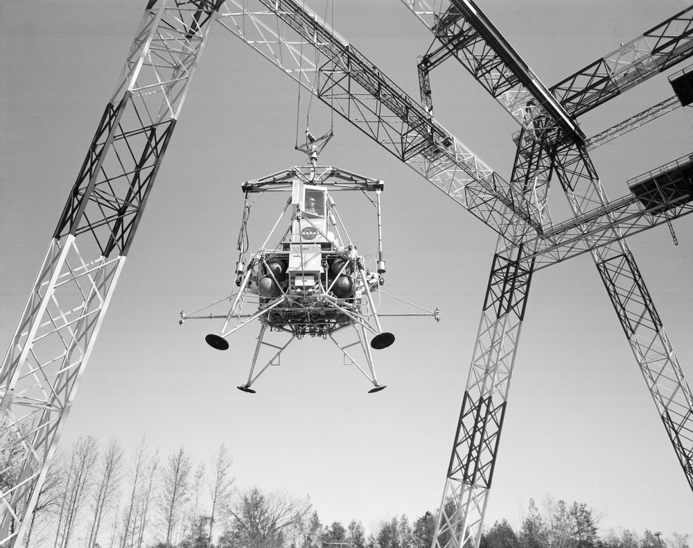 Monochrome view up at a spindly four-legged craft hanging on cables between steel towers