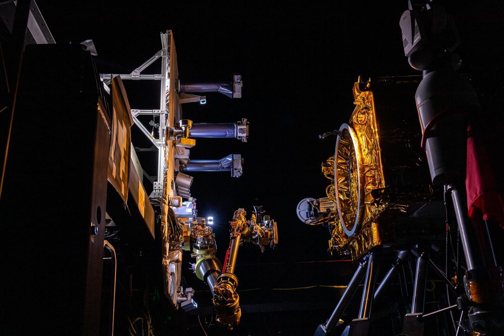 A gold-foiled satellite mock-up and a robotic arm picked out by one lamp in darkness