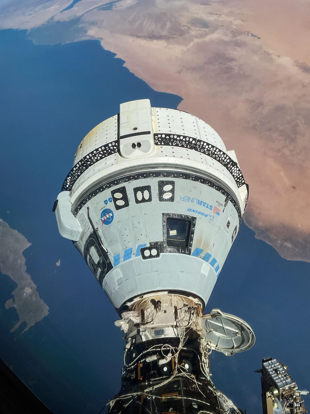 White capsule attached nose-down to the station, blue sea and desert coast below