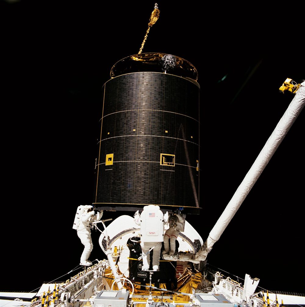 Three spacesuited astronauts bracing a tall black cylinder above the payload bay, robotic arm alongside