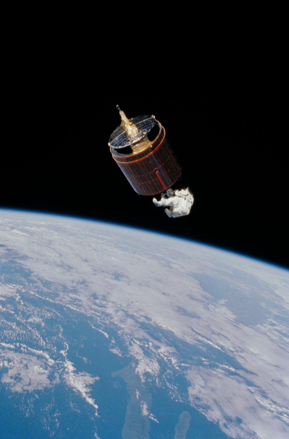 Drum-shaped satellite with a gold antenna, a white-suited figure at its base above Earth