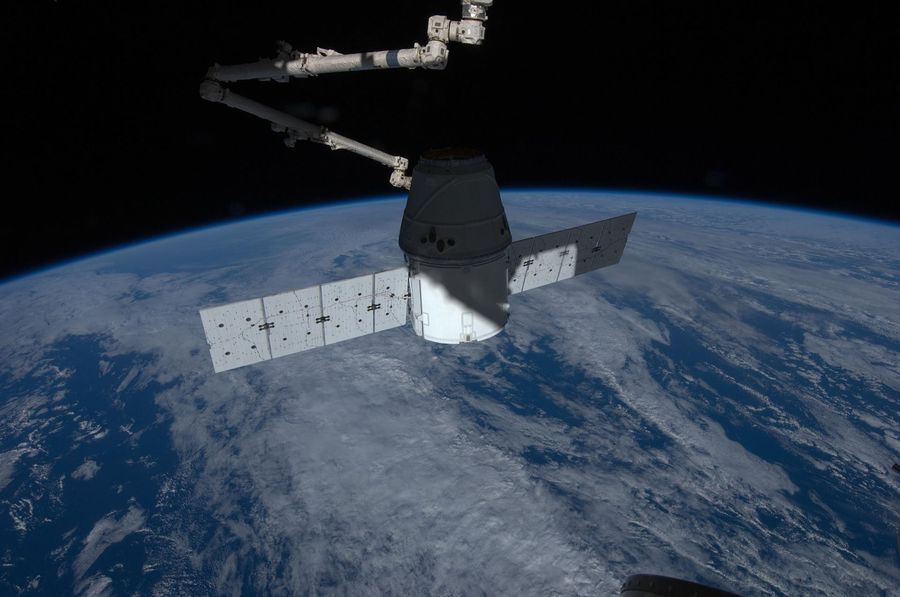 White capsule with two flat rectangular solar wings, a robotic arm reaching down towards it