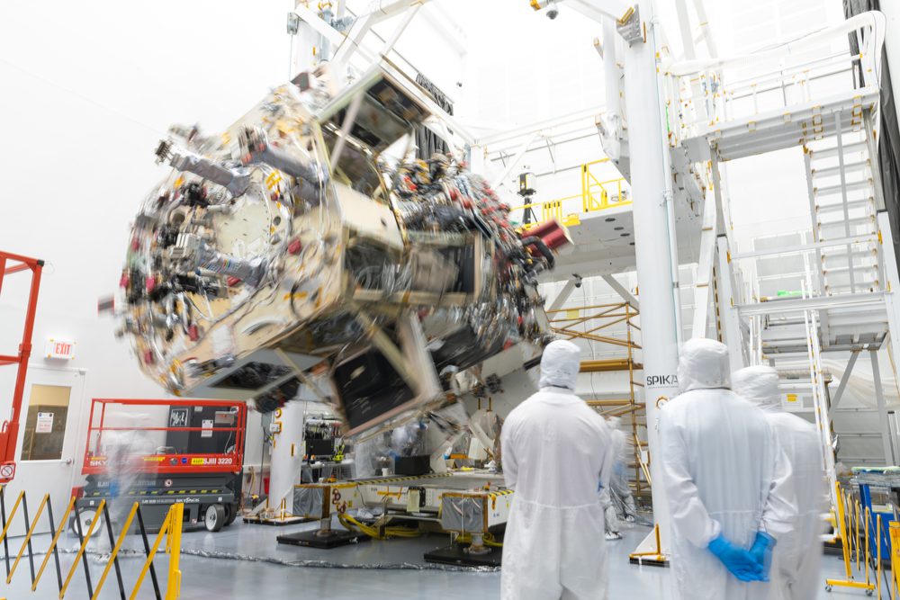 Spacecraft blurred mid-rotation on a fixture, three people in white coveralls watching from the right