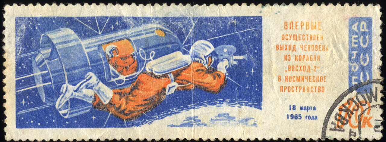 Wide blue stamp, an orange-suited figure drifting on a line from a spacecraft airlock