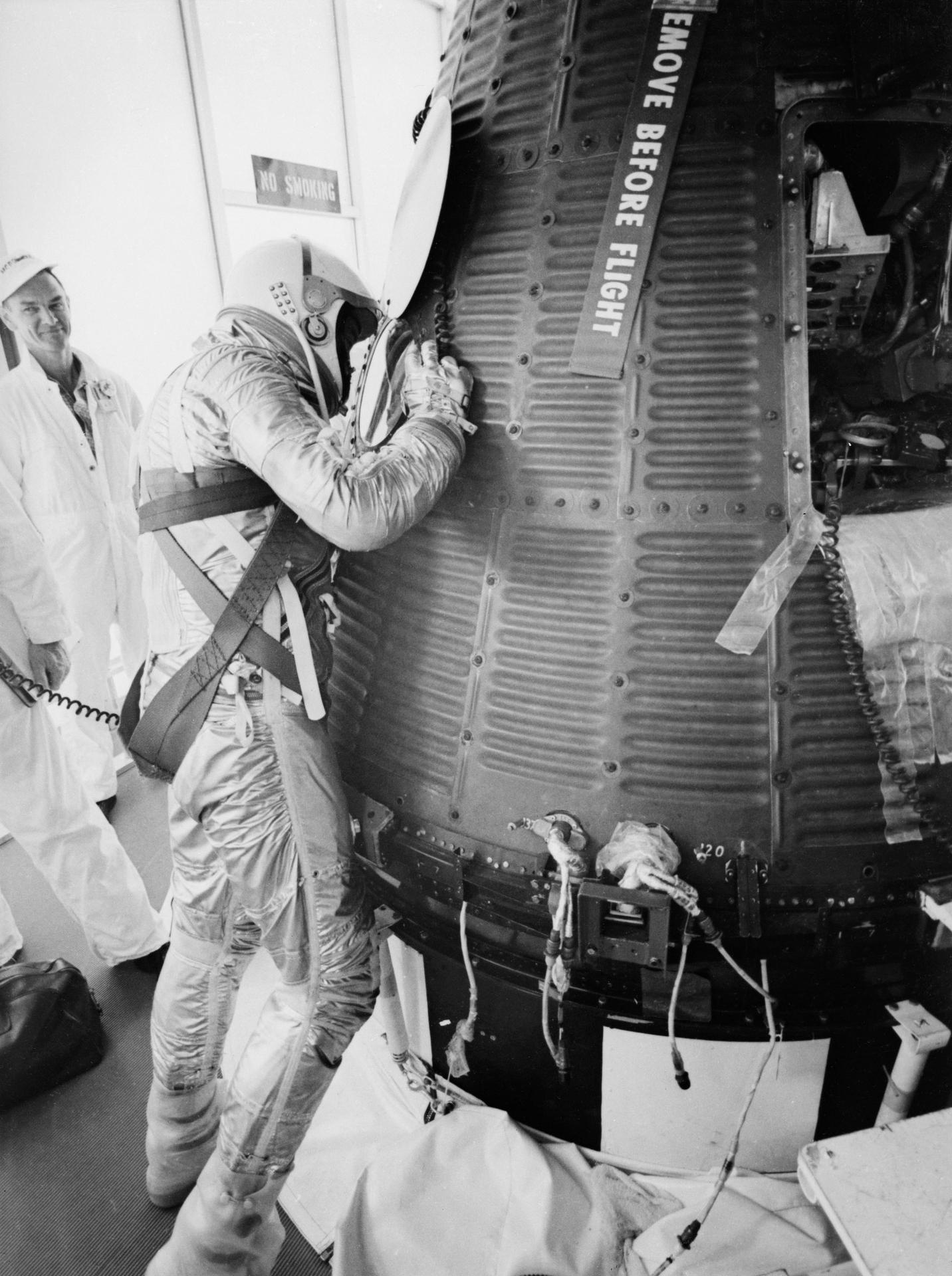 Man in silver pressure suit and helmet leaning into a small capsule hatch, technician watching