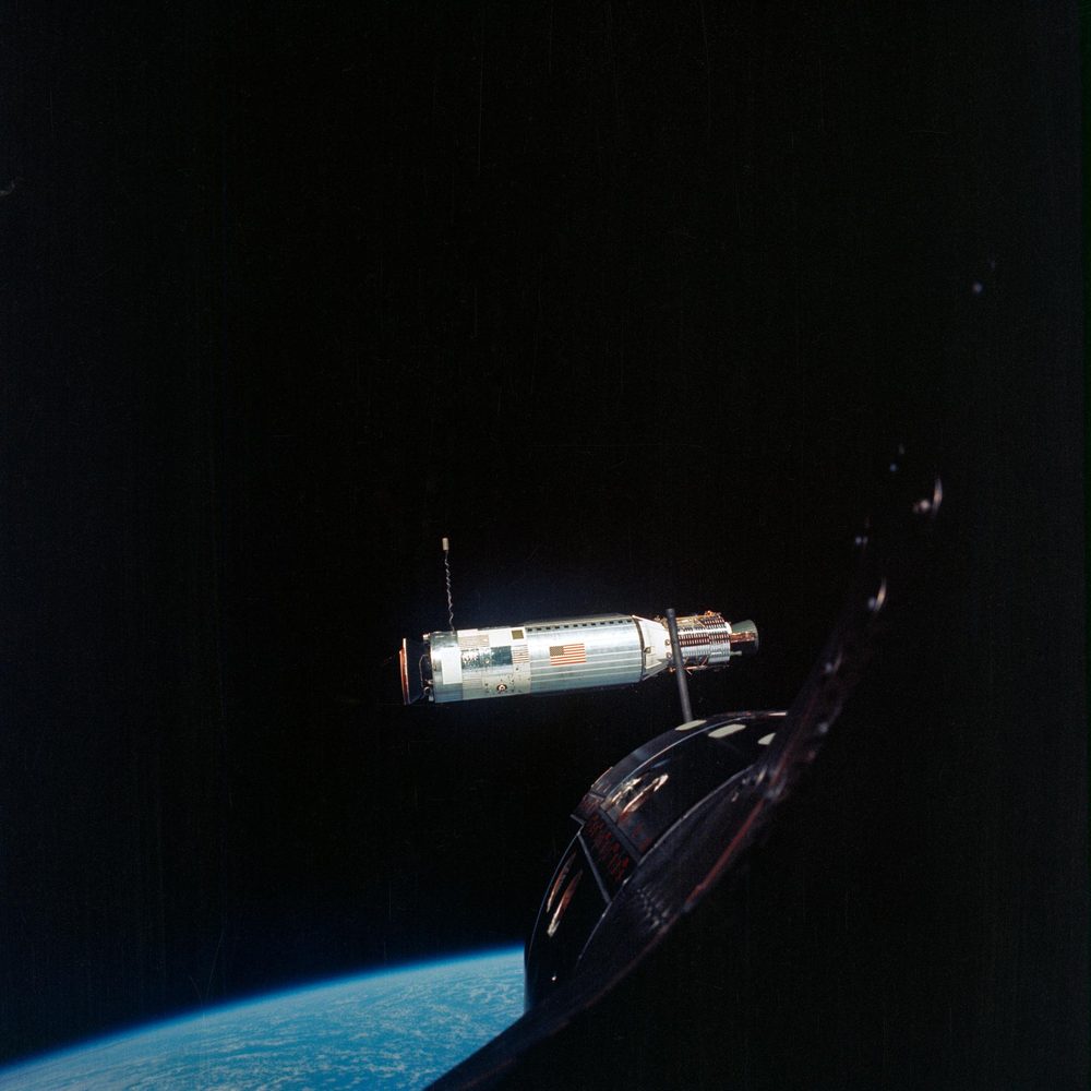 A sunlit cylinder marked with a US flag in black space, dark spacecraft nose below