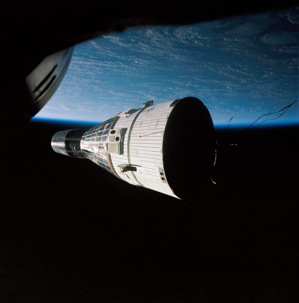 A white conical spacecraft against the blue Earth limb, seen past a window edge