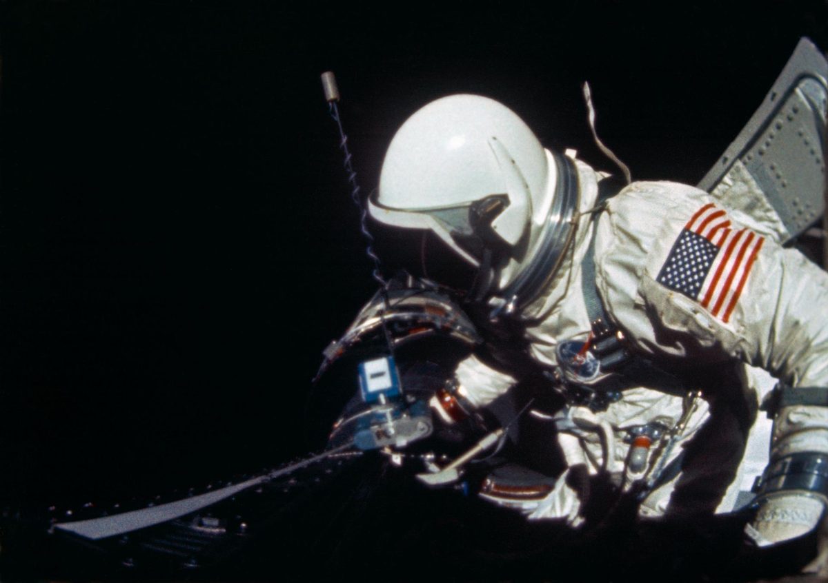 A suited astronaut leans over equipment at an open hatch, black space behind him