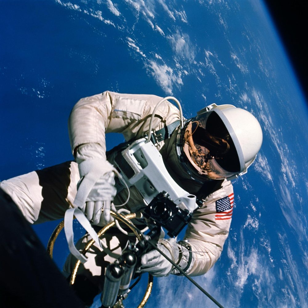 A white-suited figure floats holding a gold-tubed gas gun, gold visor reflecting the spacecraft