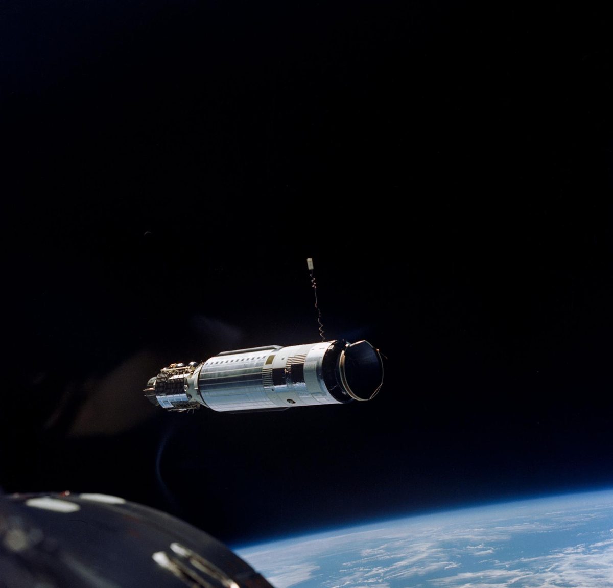 A slender cylindrical vehicle, docking cone towards the camera, above a cloud-covered Earth