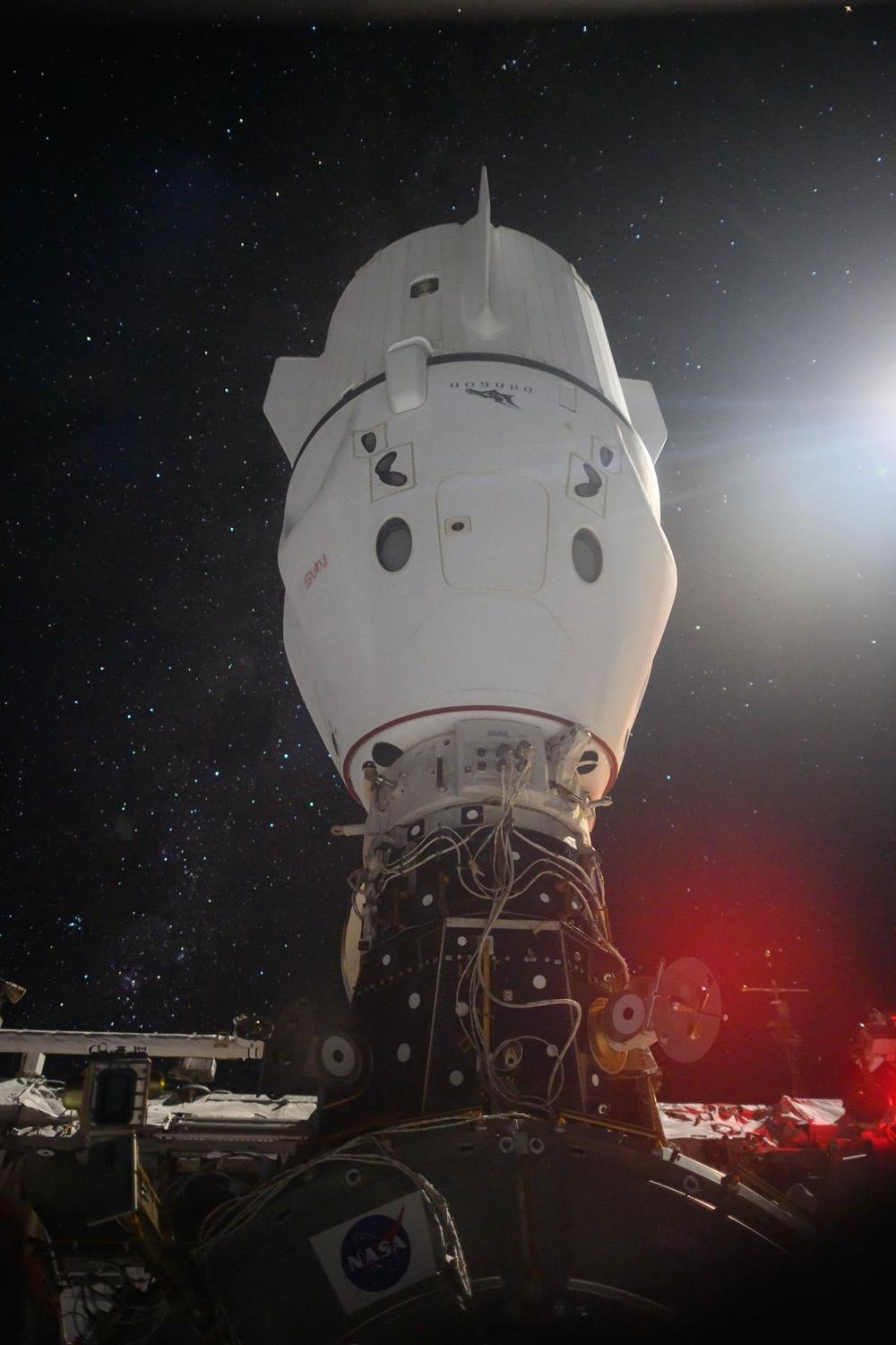 White capsule attached nose-up to station hardware against a dense field of stars