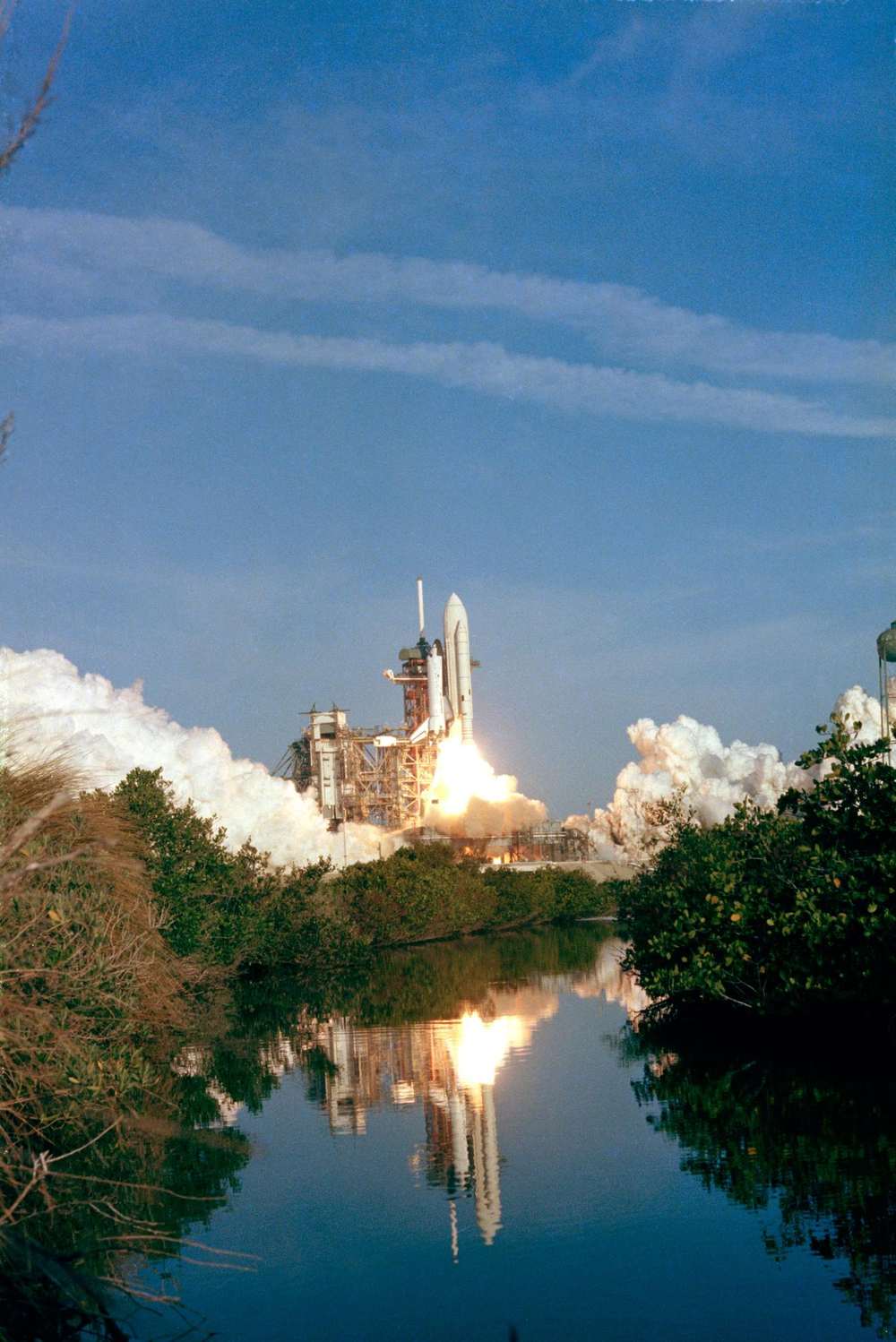 Shuttle lifting off behind low scrub, flame and steam mirrored in still water