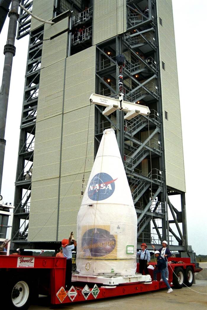 White conical shroud with a NASA insignia on a red trailer beneath a launch gantry