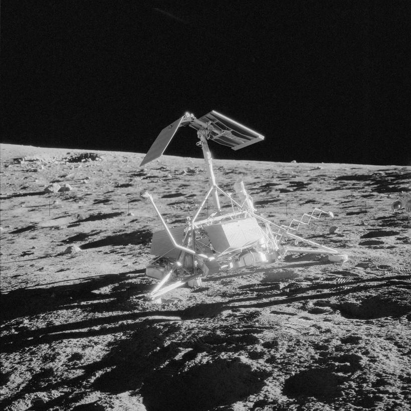 Spindly three-legged lander with a raised solar panel on grey ground, black sky behind