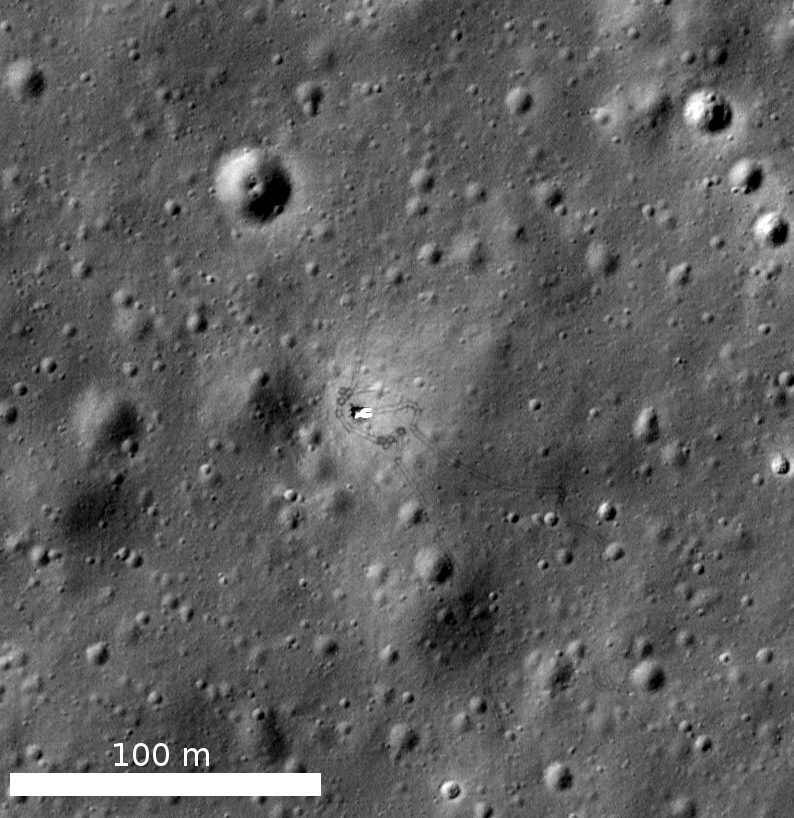 Overhead greyscale of cratered ground with faint looping lines and a 100 m scale bar
