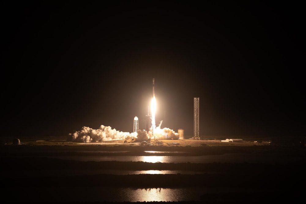 Night launch: a bright engine plume lights the pad, reflected in water in the foreground