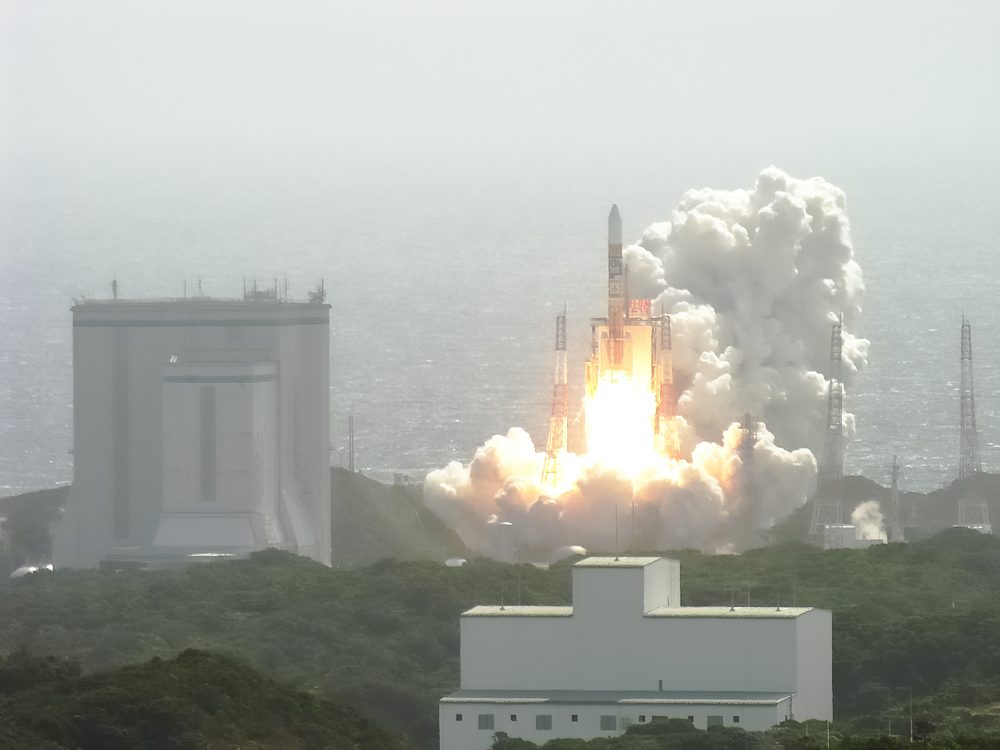 White rocket climbing on an orange flame above green coastal scrub, hazy sea behind