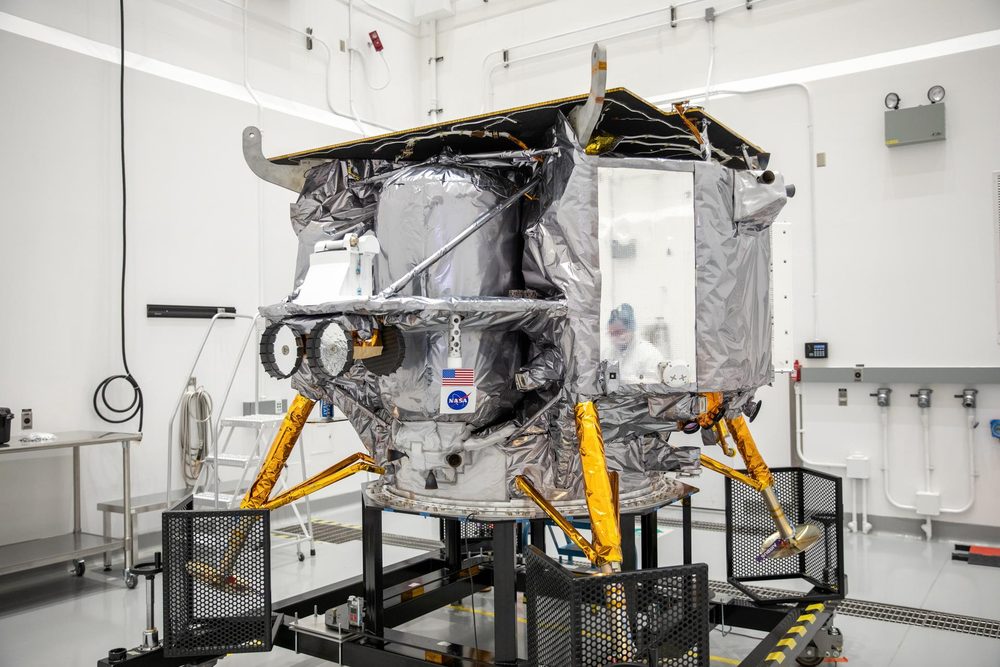 Silver-wrapped lander with orange legs on a stand in a white clean room