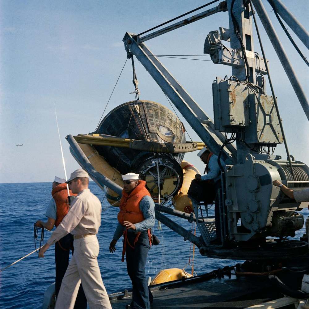 Scorched capsule slung from a shipboard crane over open sea, sailors handling lines