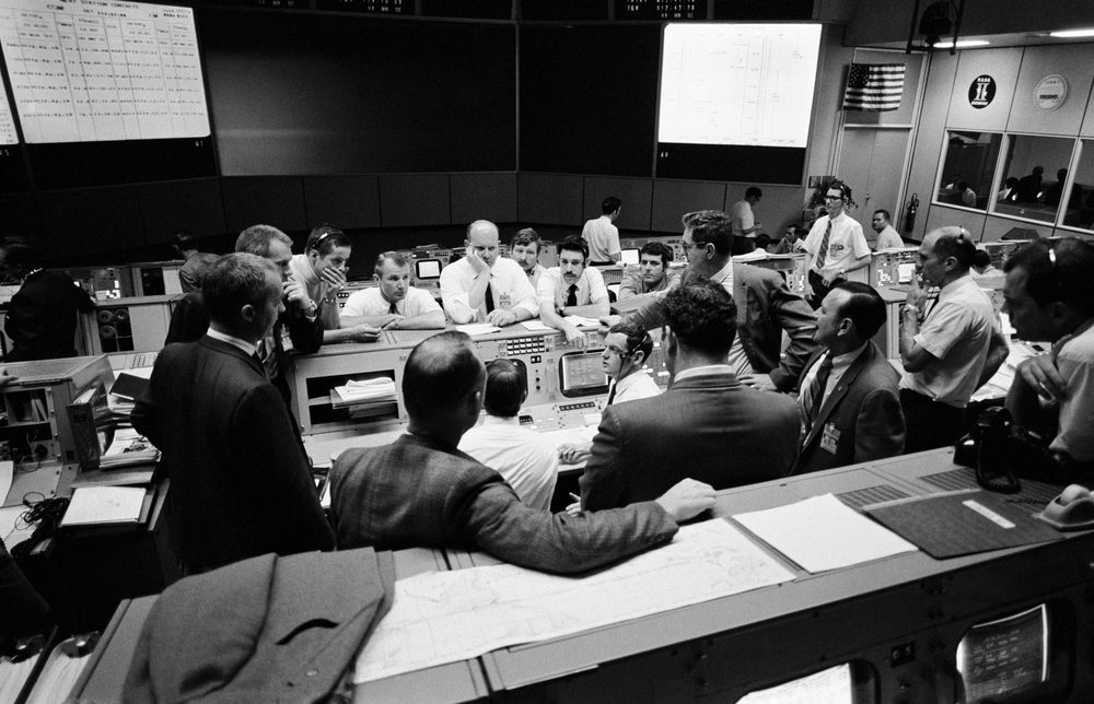 Black and white control room, men in shirtsleeves clustered at consoles beneath large screens