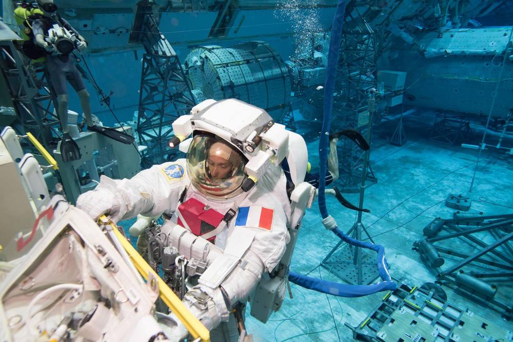A suited astronaut reaches towards pale hardware underwater while a diver films above