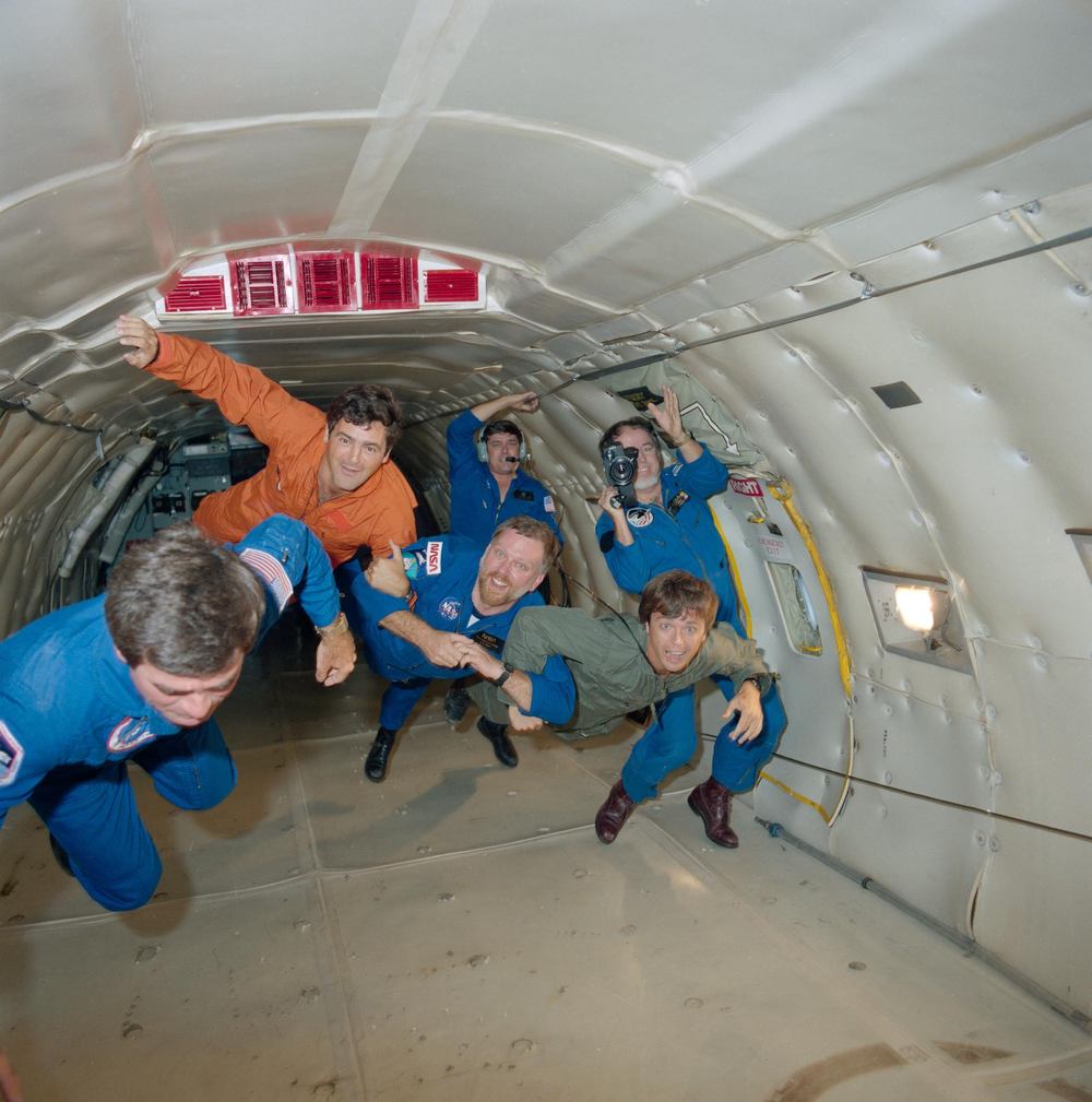 Six people in flight suits drift together down a padded aircraft cabin, one filming