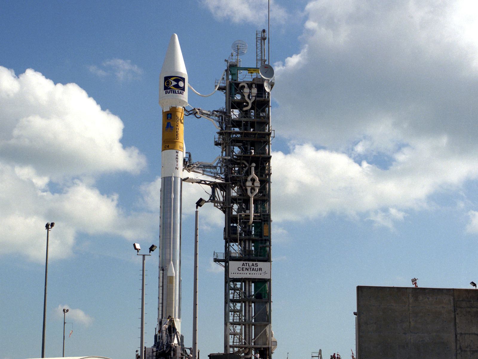 White rocket with a Eutelsat logo on the fairing standing beside its service tower under scattered cloud