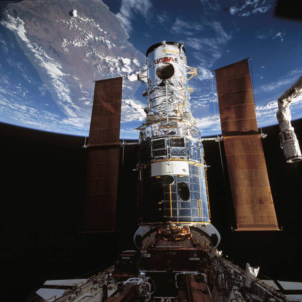 Silver cylindrical telescope upright in an open bay, two brown solar wings extended, Earth behind