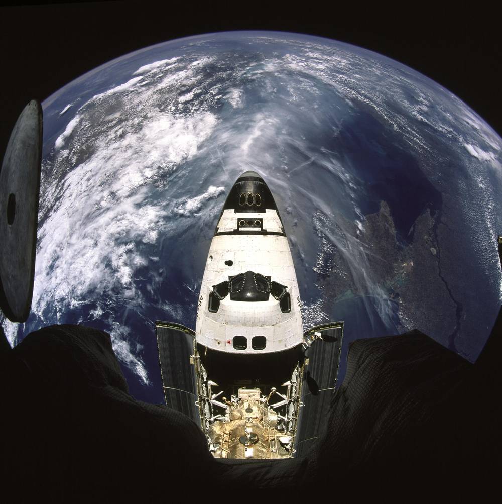 Fisheye view down onto an orbiter&rsquo;s nose and open payload bay, Earth curving behind