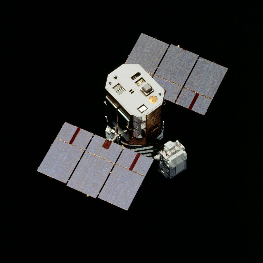 Octagonal satellite with two three-panel solar wings, a suited figure in a white backpack alongside