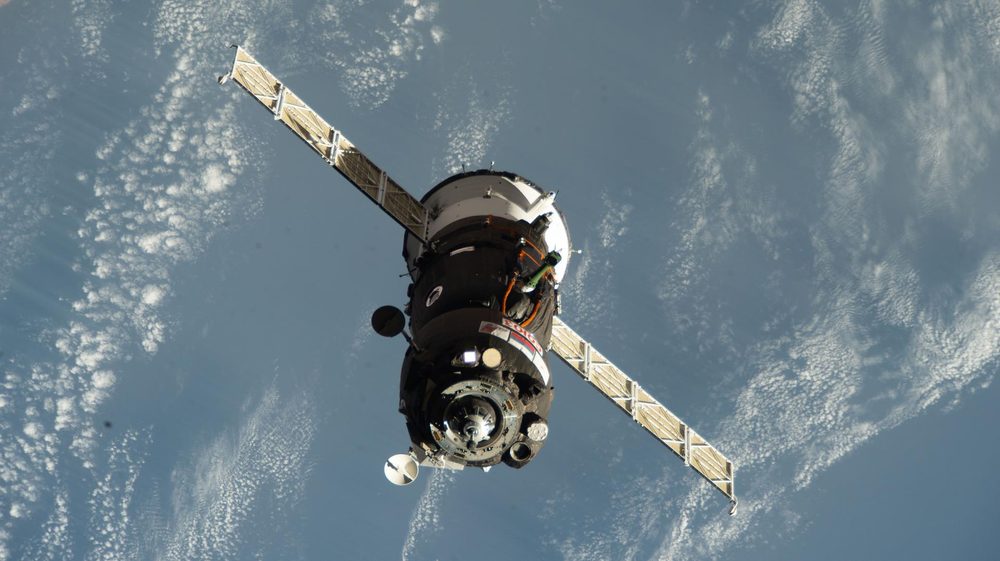 Soyuz seen head-on, two long solar wings spread, cloud-flecked ocean far below