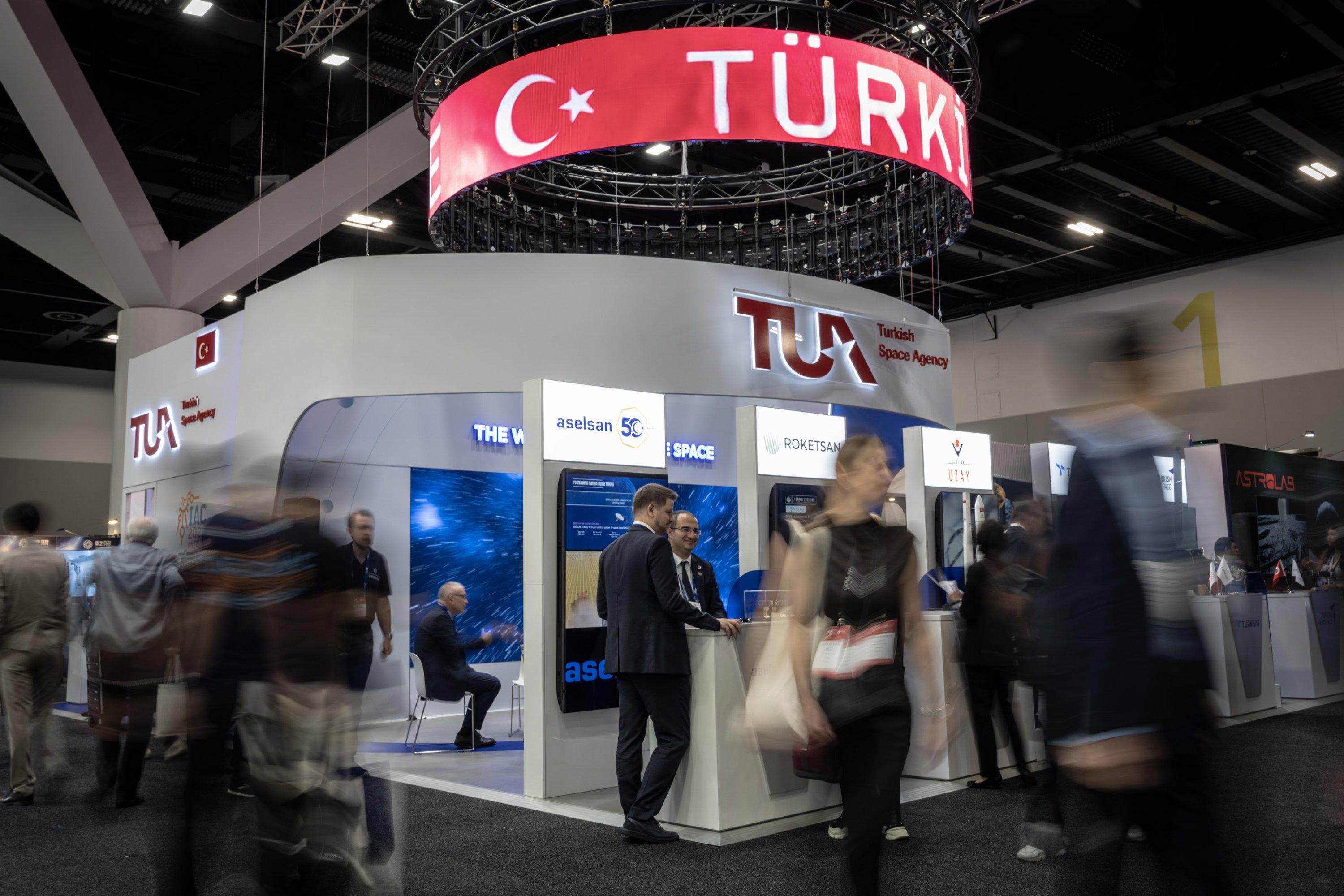 Visitors moving past the Turkish Space Agency stand at a congress, a red TÜRKİYE banner overhead and company screens along the counter